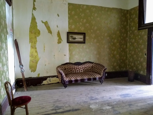 An empty room with peeling floral wallpaper, a vintage couch, and a wooden floor. A chair is in the corner.