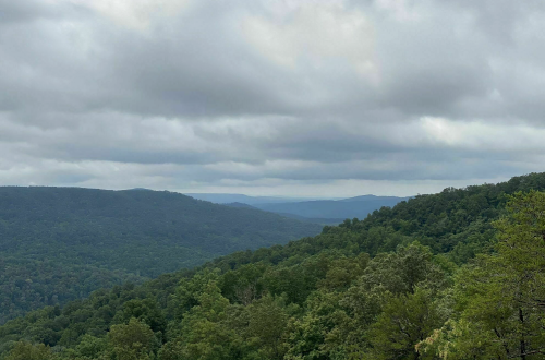 A panoramic view of rolling green hills under a cloudy sky, with layers of mountains in the distance.