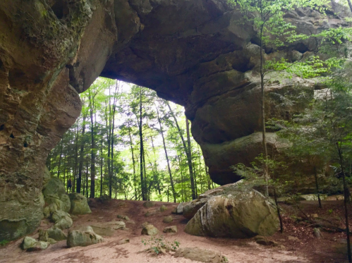 A natural rock arch surrounded by lush green trees and a sandy ground, creating a serene outdoor scene.
