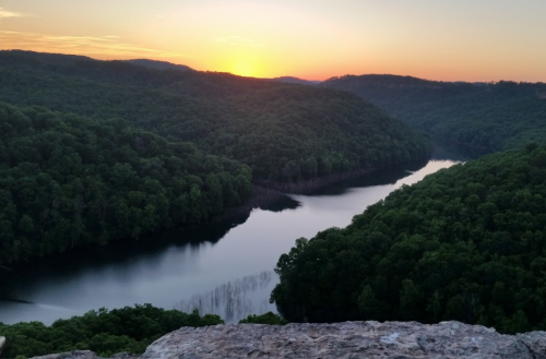A serene sunset over a winding river surrounded by lush green hills and trees.