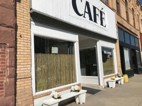 Exterior of a café with a sign, large windows covered with bamboo, and flower boxes at the entrance.