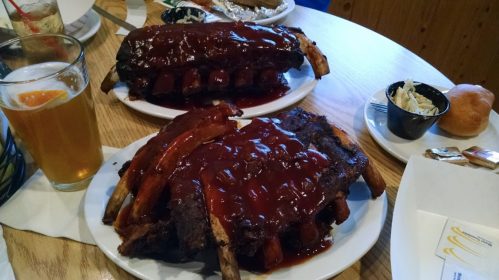 Two plates of barbecue ribs covered in sauce, served with a side of coleslaw and a glass of beer on a wooden table.