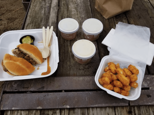 A meal featuring a sandwich with pulled meat, three cups of dessert, and a side of crispy tater tots on a wooden table.