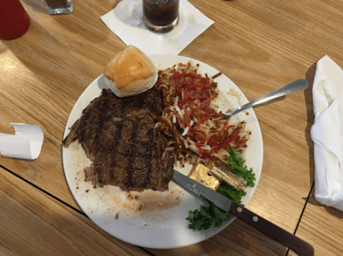A plate with a grilled steak, a dinner roll, hash browns topped with ketchup, and a side of lettuce.