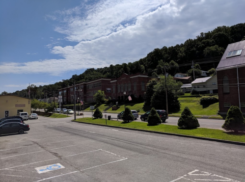 A sunny view of a small town street with brick buildings, green hills, and parked cars under a partly cloudy sky.