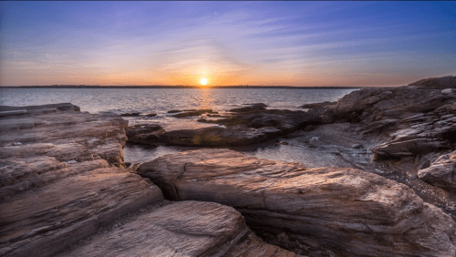 Sunset over rocky shoreline with calm water, creating a serene coastal landscape.