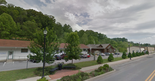 A street view of a small town with trees, parked cars, and buildings under a cloudy sky.