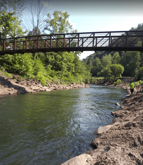 A serene river flows under a wooden bridge, surrounded by lush greenery and rocky banks. A person stands nearby.
