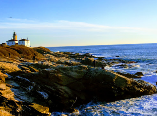 A scenic coastal view featuring a lighthouse, rocky shore, and calm ocean under a clear blue sky.