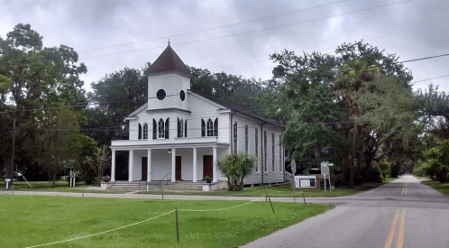 Historic white church with a steeple, surrounded by greenery, located on a quiet street under a cloudy sky.