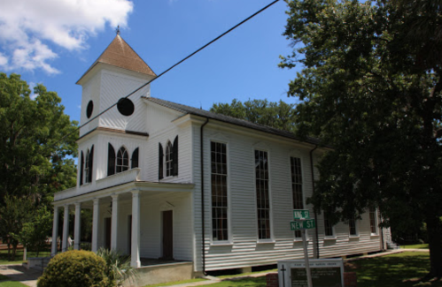 Historic white church with a steeple, large windows, and surrounded by trees under a blue sky. Street sign visible.