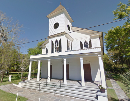 A white church with a tall steeple, columns, and steps, surrounded by greenery and trees on a sunny day.