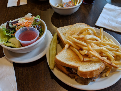 A plate with a sandwich, fries, a pickle, and a side salad with dressing on a wooden table.