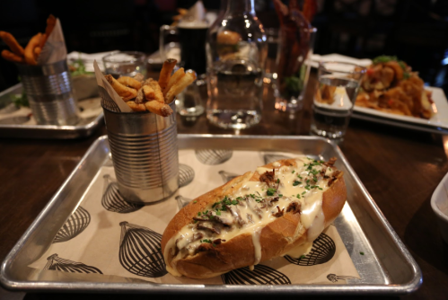 A tray with a loaded sandwich topped with sauce and herbs, alongside a cup of crispy fries in a tin can.