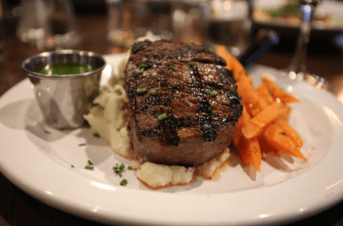 A grilled steak served with mashed potatoes, carrots, and a small cup of green sauce on a white plate.