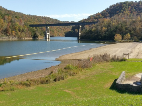 A serene lake with a bridge overhead, surrounded by autumn-colored trees and a sandy shore.
