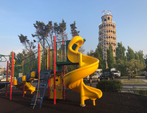 Colorful playground equipment with a yellow slide, set against a backdrop of a tower resembling the Leaning Tower of Pisa.