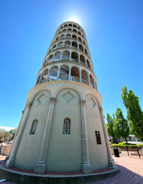 A tall, white tower with multiple levels and arches, set against a clear blue sky and surrounded by green trees.