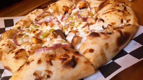 A close-up of a pizza with a golden crust, topped with cheese, chicken, red onions, and green peppers on a checkered paper.