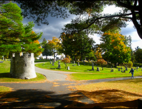 A serene cemetery scene with a stone tower, winding paths, and colorful autumn trees under a cloudy sky.