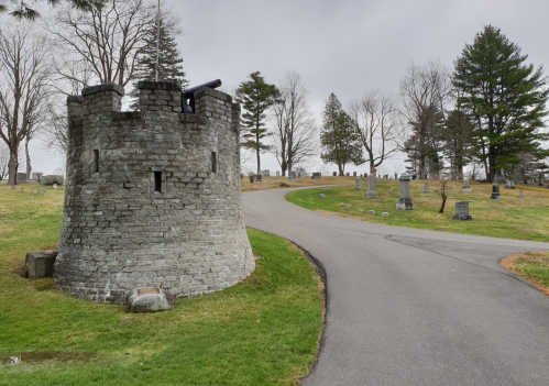 A stone tower with a cannon overlooks a winding path in a cemetery, surrounded by trees and gravestones.