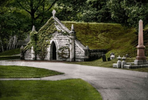 A stone mausoleum covered in ivy, surrounded by a grassy area and gravestones in a serene cemetery setting.