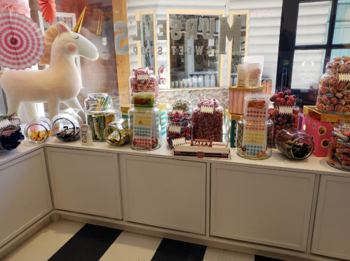 A colorful candy display featuring jars of sweets, taffy, and a large unicorn plush toy in a shop setting.