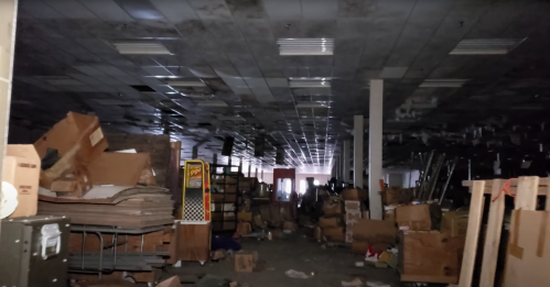 An abandoned warehouse filled with scattered boxes and debris, dimly lit with shadows across the ceiling.