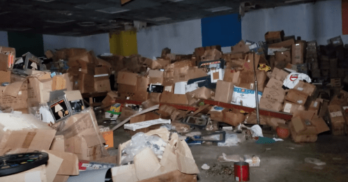 A cluttered room filled with piles of cardboard boxes and scattered debris on the floor.