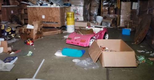 A cluttered basement with cardboard boxes and a pink toy car in the center, surrounded by various discarded items.
