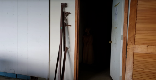 A dimly lit doorway reveals a figure standing in the shadows, with metal rods leaning against the wall nearby.