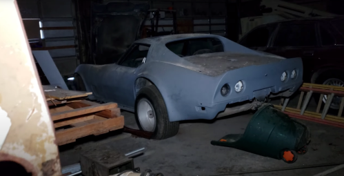 A vintage blue car is parked in a dimly lit garage, surrounded by wooden pallets and tools.