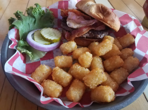 A plate with a bacon cheeseburger, lettuce, pickles, onion, and a side of crispy tater tots in a red-checkered basket.