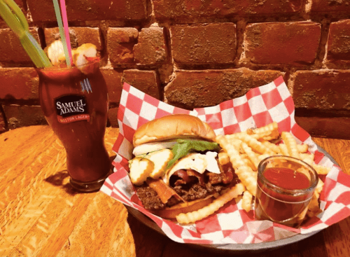 A burger with bacon and toppings, served with fries and a drink in a Samuel Adams glass, on a wooden table.