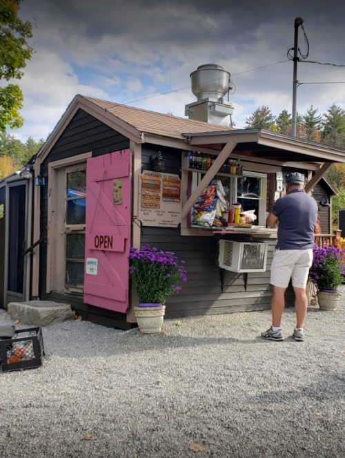 A small, rustic food stand with a pink door, open sign, and a customer ordering at the window, surrounded by flowers.