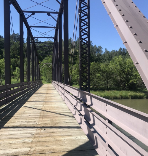 A wooden bridge with metal supports spans a calm river, surrounded by lush greenery under a clear blue sky.