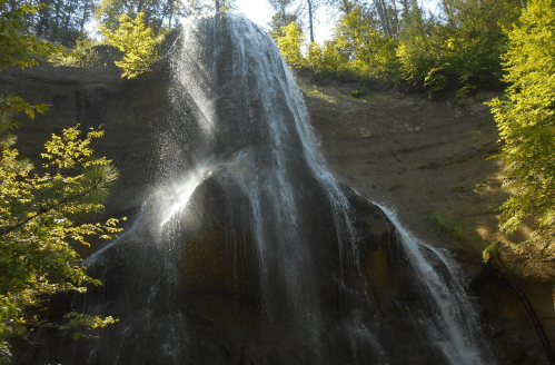 A cascading waterfall flows down a rocky cliff, surrounded by lush green trees and sunlight filtering through the leaves.