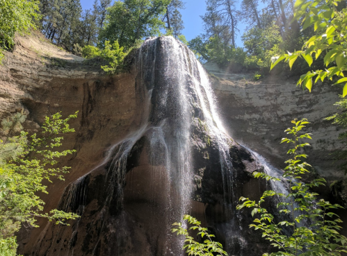 A cascading waterfall flows down a rocky cliff, surrounded by lush green trees under a clear blue sky.