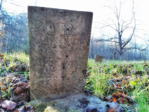 A weathered gravestone with the name "Ruby Huckaby" partially visible, surrounded by grass and fallen leaves.