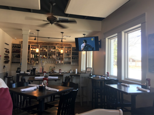 Interior of a restaurant featuring wooden tables, a bar with wine bottles, and a TV showing a sports event.