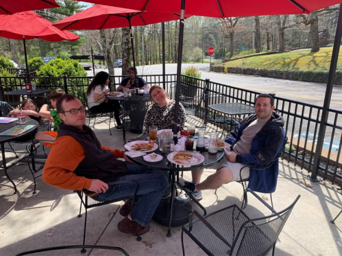 Three people sit at a table outdoors, enjoying food and drinks under red umbrellas, with others dining in the background.