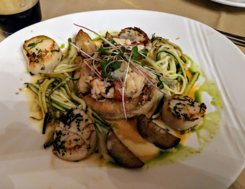 A gourmet dish featuring scallops, lobster, zucchini noodles, and garnishes on a white plate.