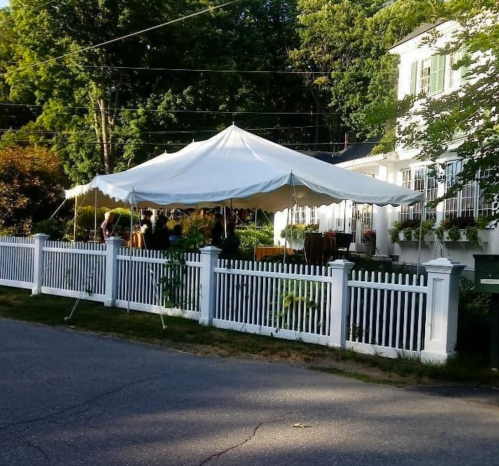 A large white tent set up in front of a house, surrounded by a white picket fence and greenery.