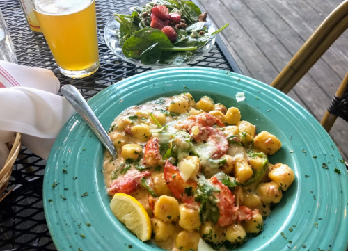A plate of creamy lobster gnocchi with herbs and a lemon slice, alongside a spinach salad and a glass of beer.