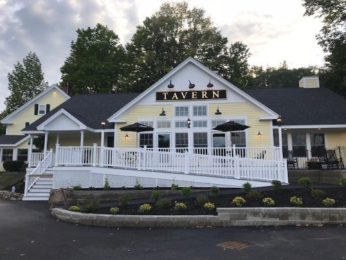 A yellow tavern building with a large sign, outdoor seating, and a landscaped entrance under a cloudy sky.