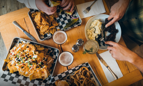 A table with various dishes: nachos, fries, steak, mashed potatoes, and drinks, with hands reaching for the food.