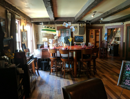 Cozy interior of a rustic bar with wooden beams, a large table, and chairs, featuring warm lighting and decor.