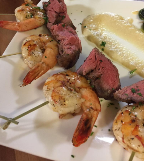 Grilled shrimp skewers and sliced beef served on a white plate, garnished with herbs and a side of creamy puree.