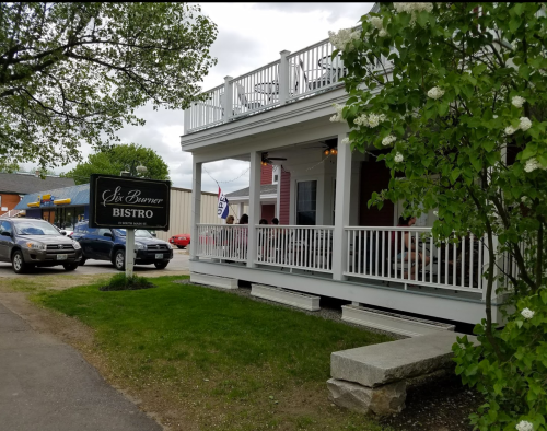 A charming bistro with a porch, surrounded by greenery and parked cars, on a cloudy day.