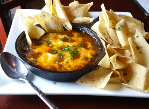 A black dish of melted cheese and beans topped with herbs, served with crispy tortilla chips on a white plate.